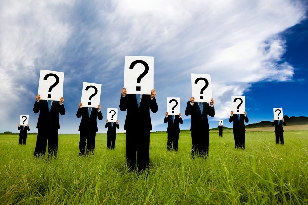 A group of people in black suits standing in a grassy field, each holding a large sign with a black question mark over their face.