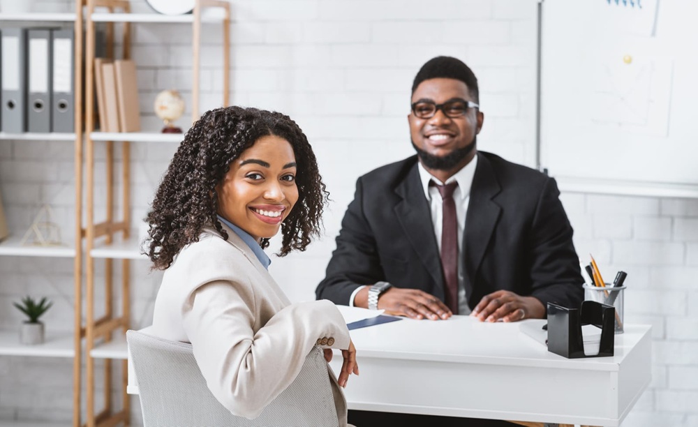 charlotte staffing companies Two professionally dressed people are sitting at a desk in an office, facing the camera and smiling. Office supplies and shelves are visible in the background.