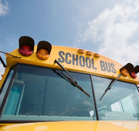 cms transportation Close-up view of the front of a yellow school bus with the words "SCHOOL BUS" on top, windshield wipers visible, and a partly cloudy sky in the background.