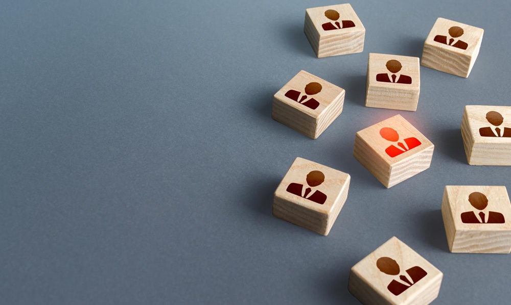 Wooden blocks with human icons are arranged on a gray surface; one block is highlighted in red, indicating emphasis or selection.
