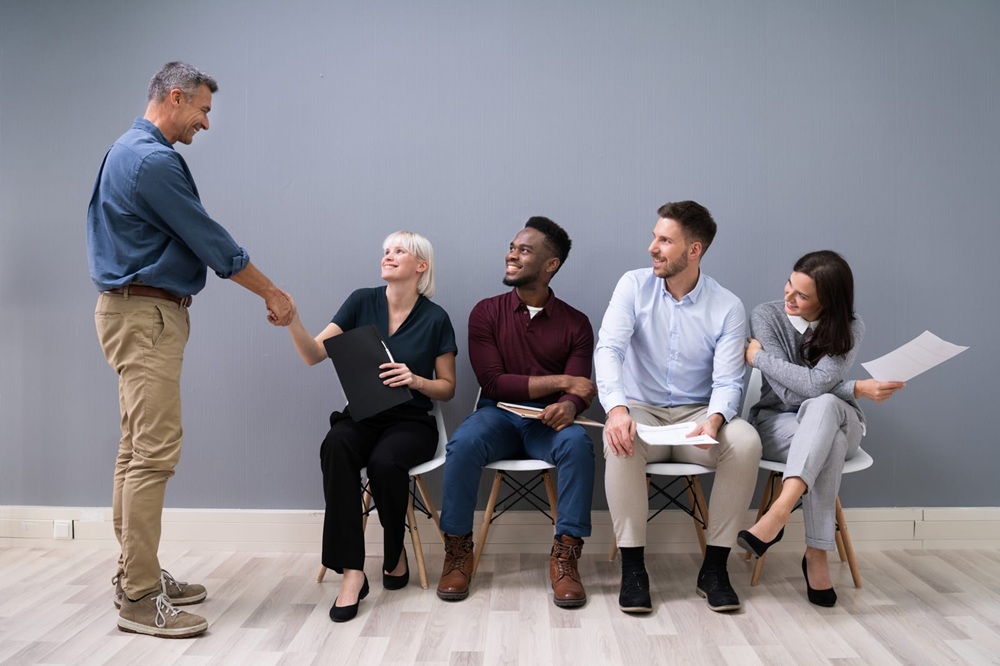 resume help charlotte nc Five people in business casual attire are in an interview setting; one standing man shakes hands with a seated woman while three others watch and hold resumes.
