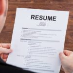 Person in a suit holding a printed resume document on a wooden desk, with a pen placed to the right of the paper.