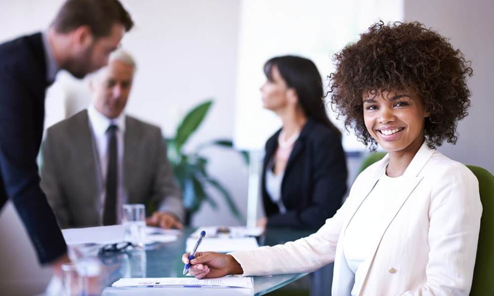 resume writing services charlotte nc A woman in a white blazer smiles at the camera while sitting at a conference table with three colleagues in a modern office setting.